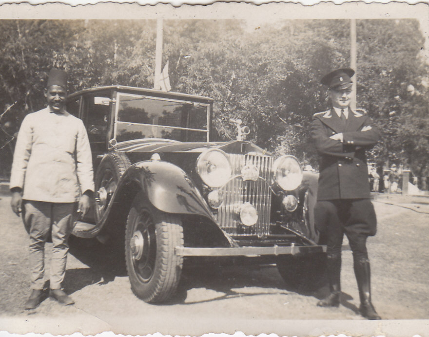 sepia photo\' of an old-fashioned black Rolls-Royce, seen almost head-on in front of trees, with two men standing buy it: on the left an Asian man in a white jacket and a fez, on the right a white man in a dark chauffeur\'s uniform and with his arms folded