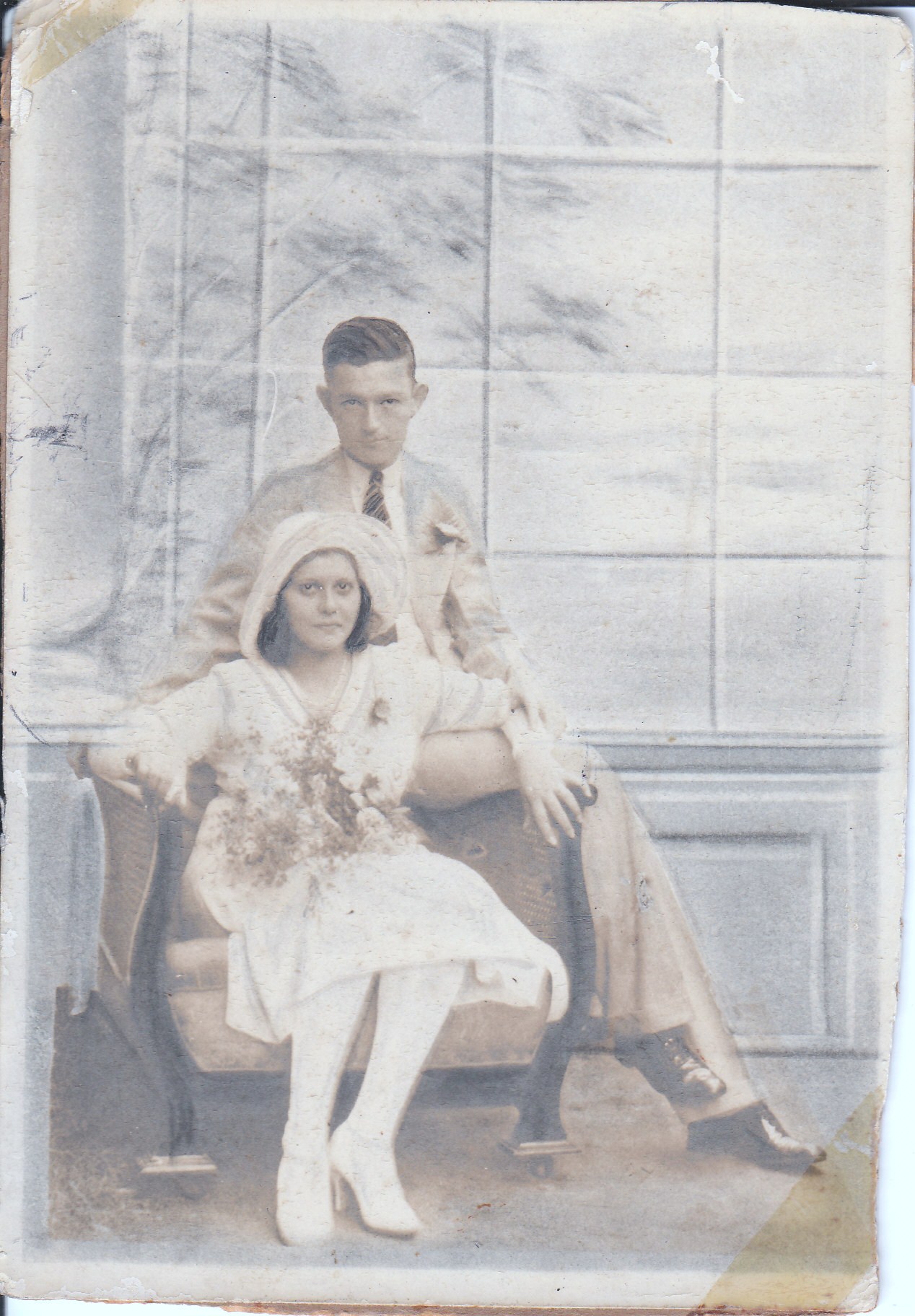 pale, faded semi-coloured photo\' of a woman in white, including a knee-length dress and big floppy hat, holding a bouquet and seated in a wide Oriental armchair, with a young man in a light-coloured suit perched on the arm on her left, and a big window or screen divided up by thin bars behind them