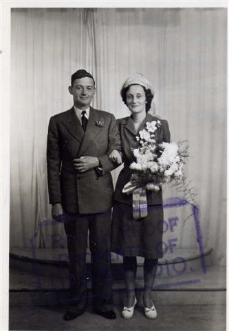 black and white photo\' of a youngish man in a mid-coloured suit, with a woman standing at his left with her arm through his, also wearing a mid-toned suit with white shoes and hat, carrying a bouquet, in front of a curtain