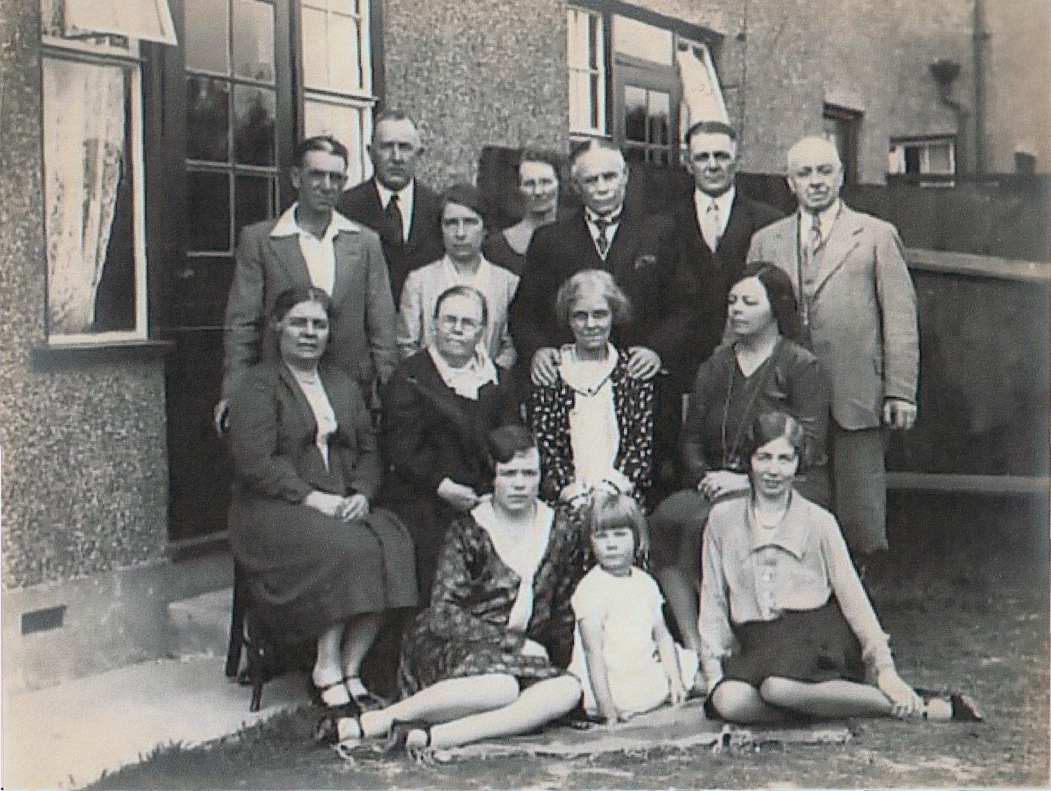 mixed group of men, women and children in 1930s clothing, ranging from about four years old to very old, formally posed in a suburban-looking garden with a standing row, a row sitting on chairs in front of them and then a row sitting on the grass