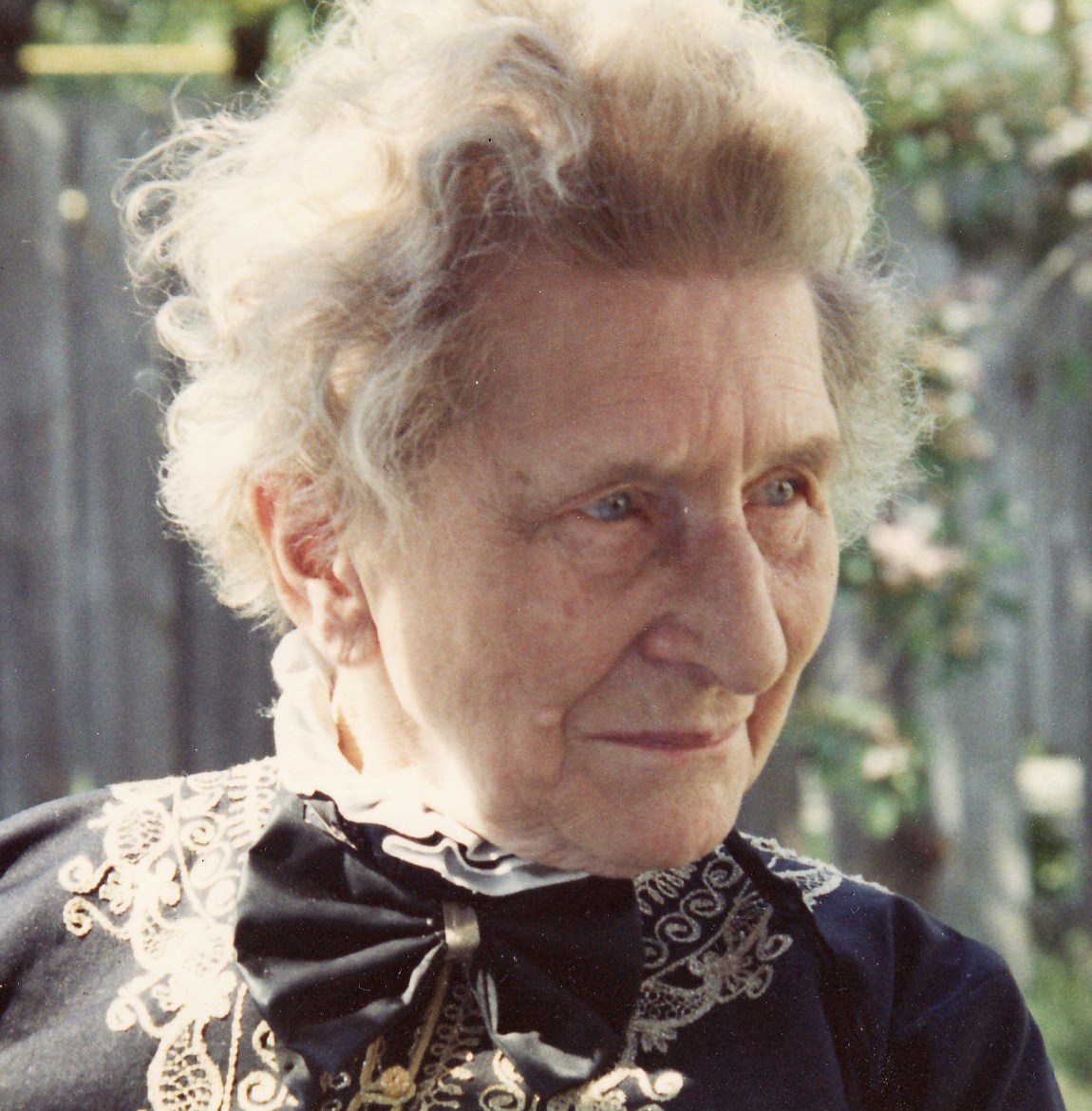 elderly lady with wavy grey hair and piercing light-blue eyes, with her head turned to look towards the right of the shot, and wearing a black blouse or dres with a cream lace collar and a black bow at the throat