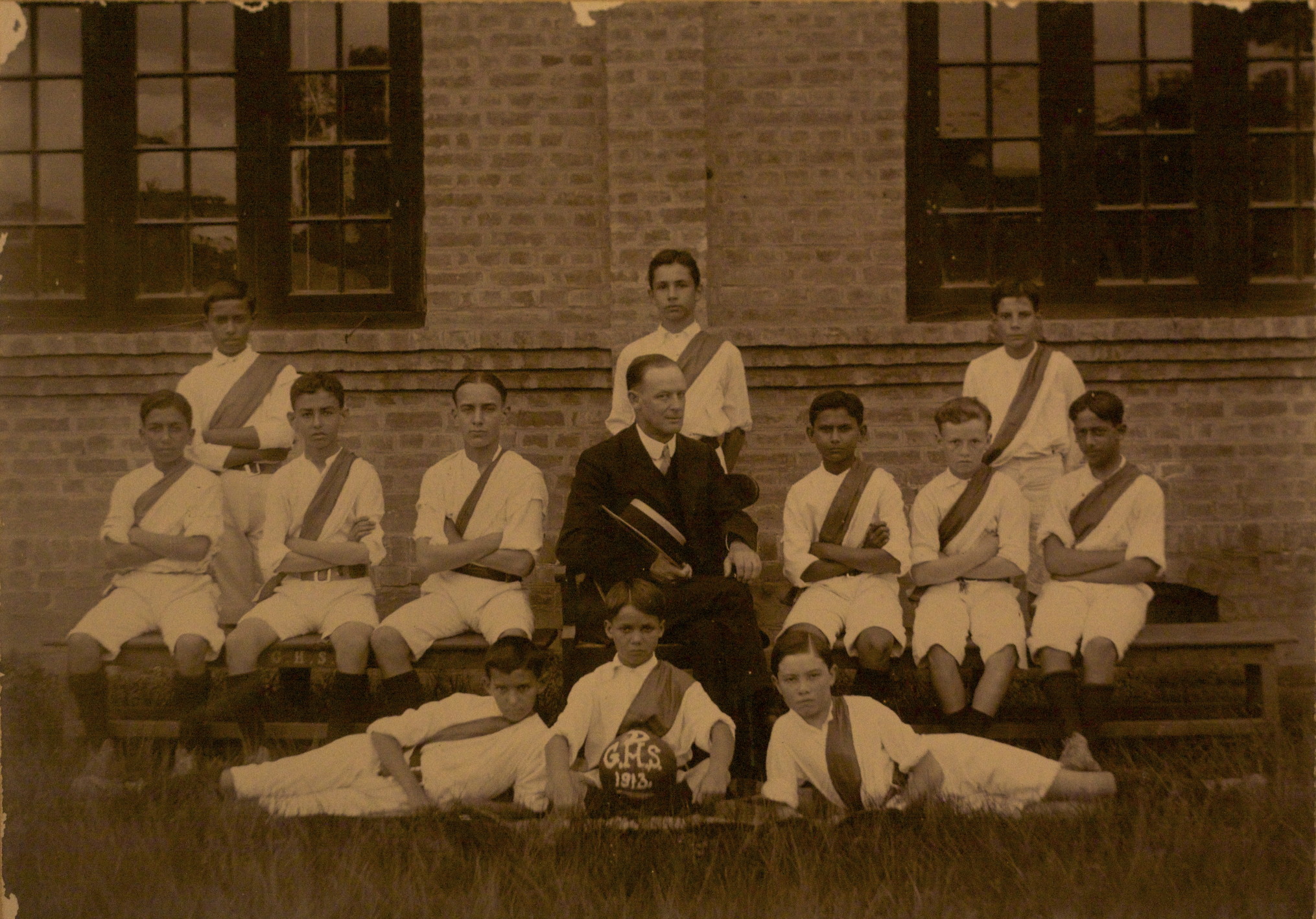 sepia photo\' showing twelve boys in white shirts and shorts with diagonal, coloured sashes across their chests, and a teacher in a dark suit and holding a straw hat, arranged in front of a brick wall with three boys standing at the back, six in front of them sitting on a bench with the teacher at the centre, one sitting upright on the grass holding a football and one either side of him sprawled on the grass