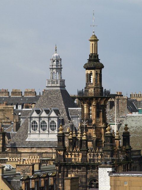 complex roofscape out of which protrude in the foreground a sort of skinny openwork pagoda in dirty cream stone, surrounded by small pillars topped with balls, and in the background a grey-tiled, four-sided pyramid with an angular triple dormer on each side, and topped with a silvery lantern
