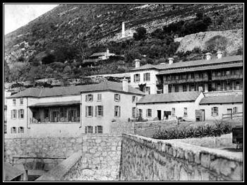 black and white photo\' showing cluster of three- or four-storey white buildings with low stone walls in front of them and the ground rising steeply behind