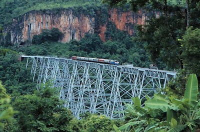 diagonal view across a valley filled with dense jungle looking towards bare reddish cliffs in the background, the foreground crossed by a narrow bridge supported on flimsy white-painted metal pylons which look like the Eiffel Tower made from thin wire, with a train crawling across the top