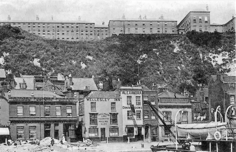 black and white photograph showing old-fashioned shops lining a quay with a couple of small boats drawn up out of the water in the foreground, while behind the shops is a medium-height white chalk cliff mostly covered with foliage, and crowned with a high, long, rather bleak-looking multi-storey municipal building which adds substantially to the height of the cliff