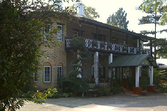 long two-storey building in snuff-coloured brick, set among trees and viewed from a corner which bulges out into a round bay: the tall oblong windows are divided into small panes and decorated with brick arches over the top, and along the front of the building, facing to the right, there is a full-length black and white balcony supported on white pillars, and a projecting porch also with white pillars, which are seated into red blocks at the foot