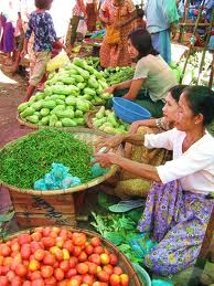 Asian women wearing pale blouses and long, brightly-coloured skirts, sitting on the ground by huge baskets of fresh fruit, vegetables and spices