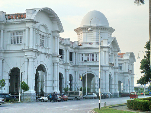 angled shot across a large two-storey white building, near the viewer at the left of the frame and far away at the right, having columns, colonnades and domes along the front, done in a square, chunky style