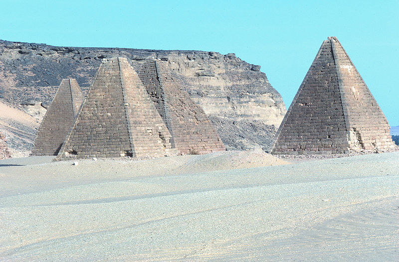 group of small, elongated pyramids in a painfully-bright desert landscape with a low cliff behind them