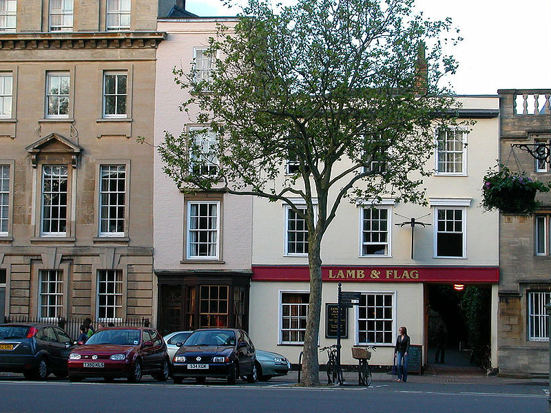 flat-fronted off-white Queen Anne-style pub with the name \'Lamb & Flag\' painted on a dark-red bar of colour between the ground and first floors, with a tree in front and flanked by other buildings, some painted, some of grey stone