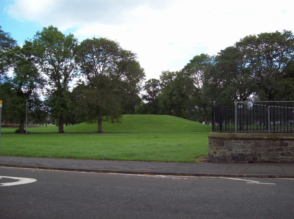 view of a large grassy mound surriounded by lawns and trees, with the edge of a school playground at right foreground