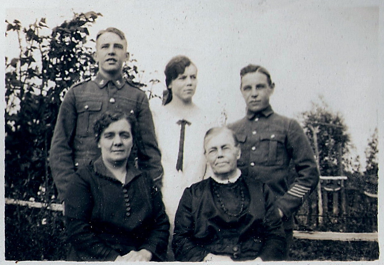 black and white photo\' showing two seated women, the one on the left middle-aged with dark hair, the one on the right elderly, both wearing black and visible only from the waist up: behind them stand a teenage girl dressed in white flanked by two boys in uniform, all set in a garden