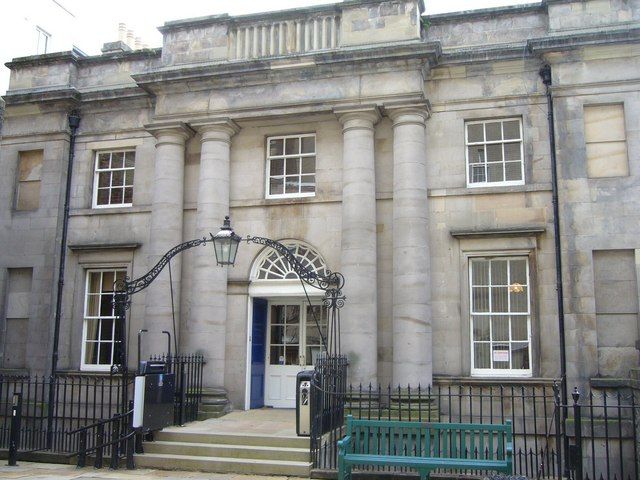 medium-sized but massive-looking two-storey grey stone building, the front door and a window above it framed by pairs of classical pillars, with a balustrade around the roof, steps up to the door with an ornamental metal arch suspending a lamp over the steps, and a green wooden bench in front of the building on the right