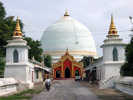 view towards a great white dome crowned with a small gold spike, along a path flanked by two white pillars also crowned with gold spikes: at the foot of the dome is a small, elaborate red and gold building