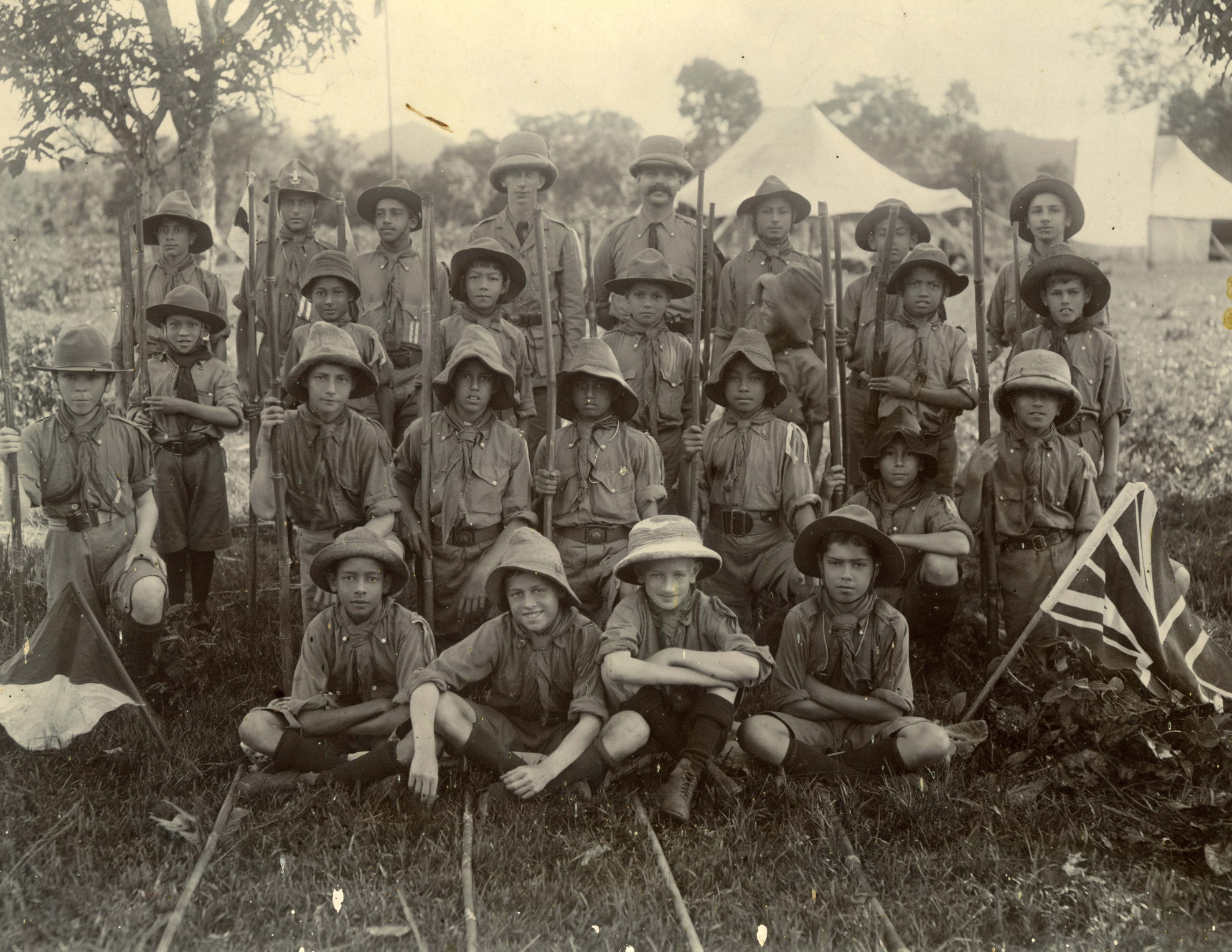 greyscale photo\' showing twenty-four boys and two young men, all in scout uniform with their hats on, lined up in four rows with white tents behind them and flags displayed on either side