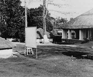black and white photo\' of a whiteboard set up on a lawn which is surrounded by trees, a low building with a white-pillared verandah and some kind of thatched outbuilding
