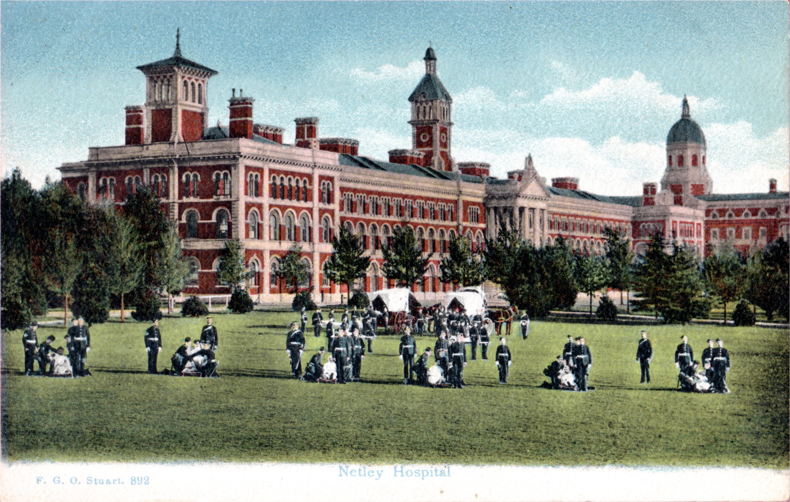postcard showing a tinted photo\' of a large, long three-storey red and cream Victorian building with twiddly turrets, with a formation of men in dark uniforms and patients in wheelchairs drawn up on a front lawn dotted with trees