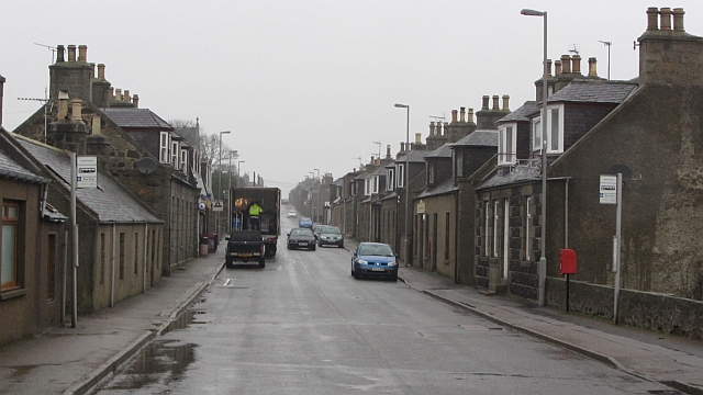 view down a wide, wet street lined on either side with small, square, grey stone houses with dormer windows