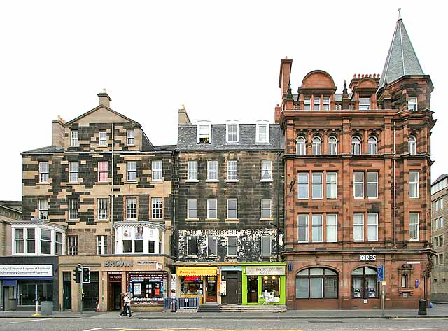 modern photograph of a row of stone-built Victorian shop-fronts: on the right, on the corner of a side-road, is a tall brown building crowned with a spire