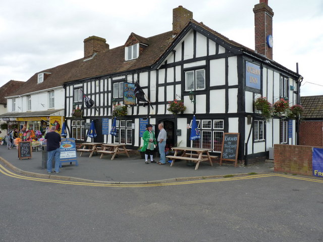 row of very old-looking shops, terminating in the right foreground with a two-storey black-and-white timber-framed pub hung about with hanging baskets of flowers: in front of the pub are some A-frame bench-and-table sets and two men one of whom is wearing a green Seventeenth Century costume