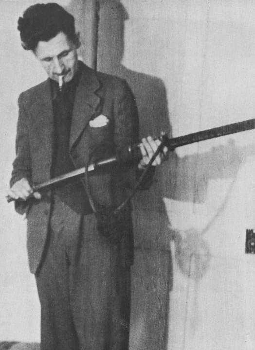 greyscale photo\' of a tall cadaverous middle-aged man in a dark suit, with a wild bush of black hair and with a cigarette dangling from his lips, standing looking down at an Oriental sword which he is holding horizontally and has half drawn from its scabbard