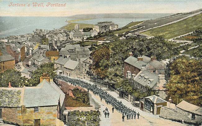 old, tinted aerial postcard photograph of a village with the sea in the background, and a column of soldiers marching along the main street