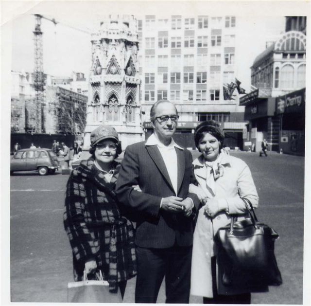 black and white photo\' of a middle-aged man between two women, one on the left in a loud check or tartan jacket, one on the right in a fawn mac, all standing in front of the Eleanor Cross at Charing Cross