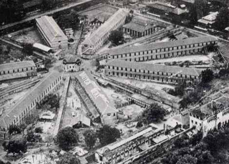 black and white aerial view looking across a group of eight long two-storey barracks-blocks radiating from a central hub, with a large high entrance-building at bottom right