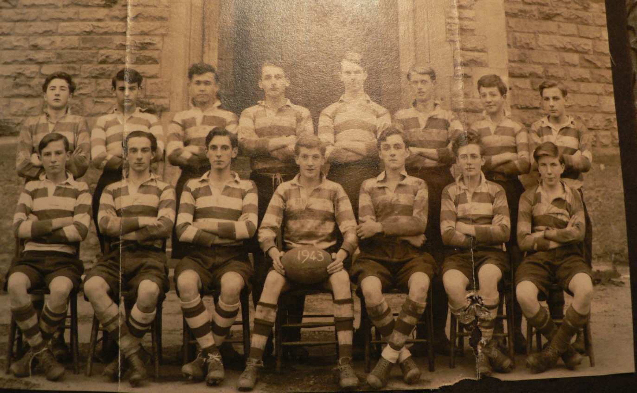 sepia photo\' of eight standing and seven seated teenage boys wearing sports outfits including jerseys striped with wide horizontal pale and dark bands, with the middle boy at the front holding a rugby-ball with \'1943\' painted on it
