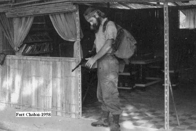 black and white photograph of a youngish, bearded man wearing combat gear, a rucksack and a headband, standing looking down at a gun in his hands, in front of a flimsy canvas and bamboo building