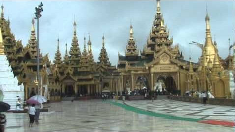 view across a rain-washed paved floor towards an arc of smallish golden pagodas with multi-tiered, spiky roofs and and pillar-framed doorways