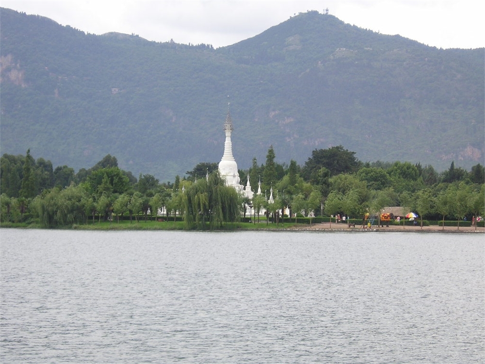 view across an expanse of water towards a white pagoda among trees, with dark, wooded mountains rising behind