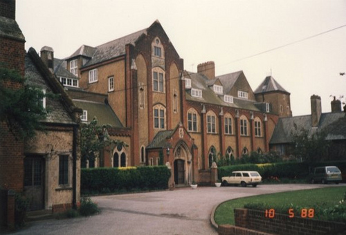 angle shot across a four-storey building in ginger brick with brown trim, with a slate roof, two of the storeys being in the roof, with a driveway and lawn in front