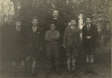 greyscale photo\' showing five boys in uniform standing in a row with a black-robed priest behind them, in front of trees