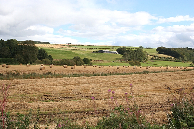 view across cut hayfields, with hay-bales, towards low rolling green hils dotted with clumps of trees, with farm buildings in a hollow at centre distance, and more hayfields crowned with dark trees at far left