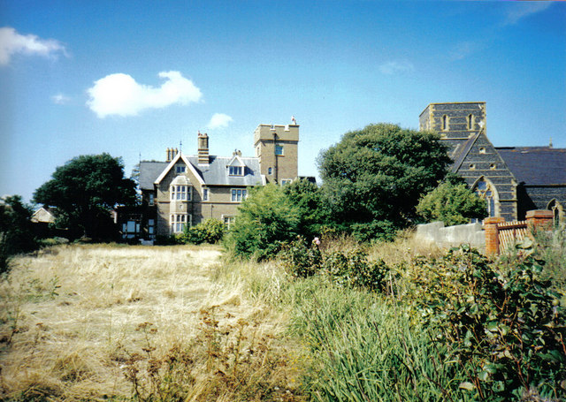 view across a rough field edged with bushes, toward a Victorian Gothic building in beige brick with a square tower on the right, and further right a large square-towered church in slate-grey brick