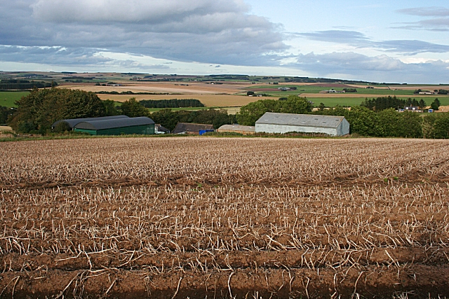 view from a hill down over a brown field towards a group of farm buildings with rolling hills in the background