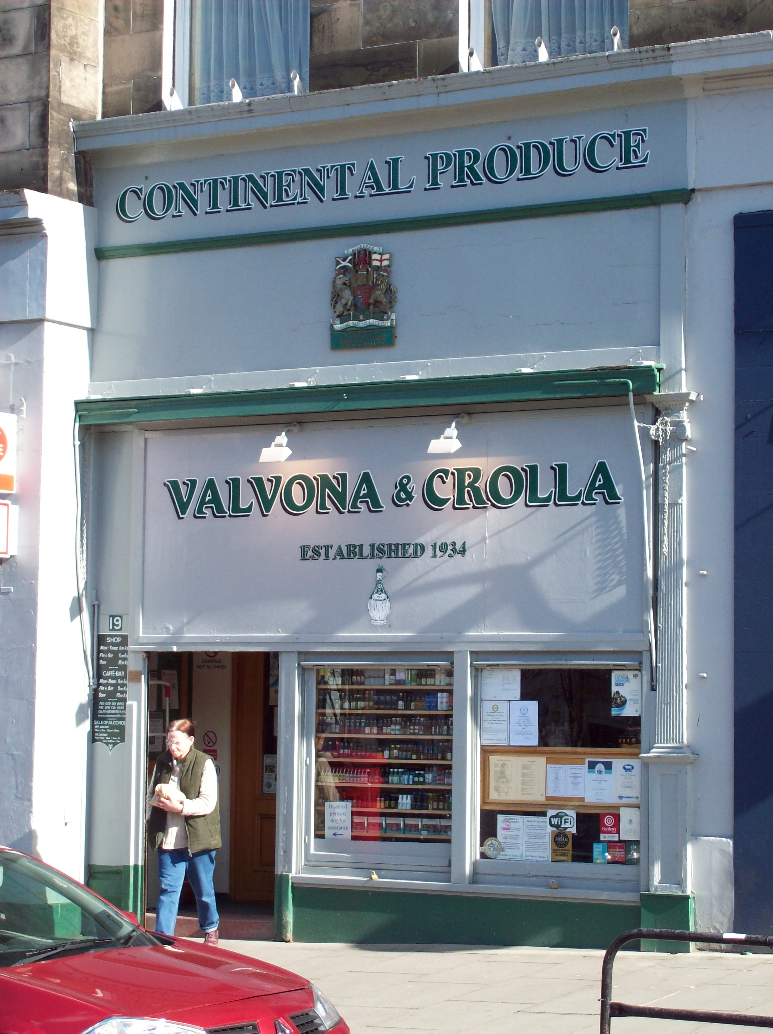 pale blue or blue-grey shopfront trimmed with grey-green, with an upper storey of brownish stone showing above; the shop window is crowded with notice-boards and shelves of miniature bottles and the pavement in front of the shop slopes noticeably down to the left