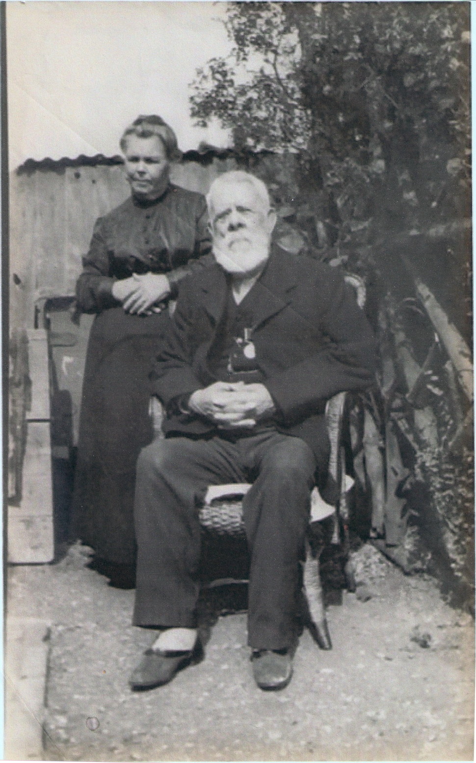 elderly, white-bearded and snub-nosed man in a dark suit, seated in a wicker chair in a garden; a middle-aged woman with blonde hair twisted up in a bun, a dark blouse and an ankle-length black skirt stands behind him on his right