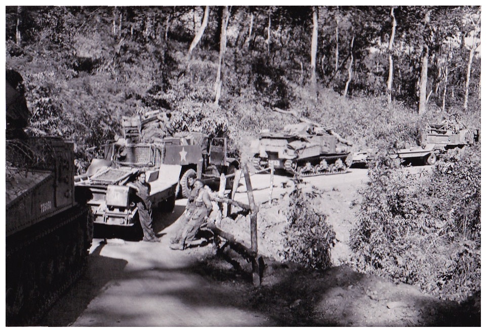 greyscale photo\' showing a mixed column of armoured cars and tanks, one with a U.S.-type white star, parked on a winding road at the foot of a wooded slope: a man is standing leaning against an armoured car, and two others are sitting on a crossbar of a fence, facing him