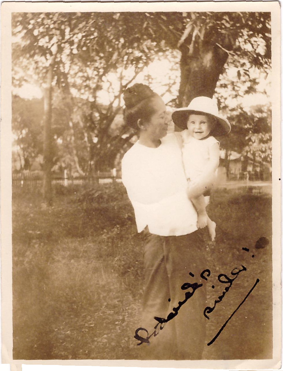 Asian lady stands under a tree, holding baby Rory who is wearing a topee and smiling broadly