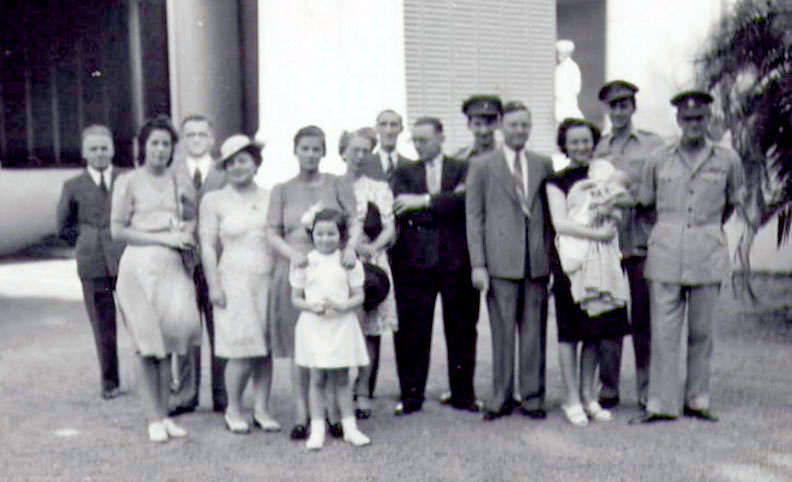 black and white photo\' of fifteen people standing in a group, including a babe in arms and, in the foreground, a little girl in a short white dress and with a white bow in her hair
