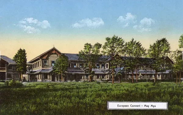 hand-coloured photo\' of long wooden two-storey building with an expanse of grass and some trees in the foreground