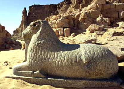 slightly stylised statue of a reclining ram, with the hill of Jebel Barkal, a very eroded standing statue and a jumble of fallen rocks and masonry in the background