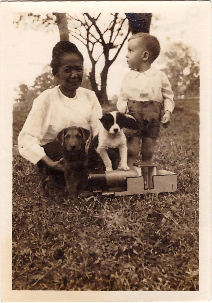 Rory, aged about three, stands in a garden looking off to his right; an Asian lady crouches next to him holding two puppies, one brown, one black and white, and there is a large toy train in front of them