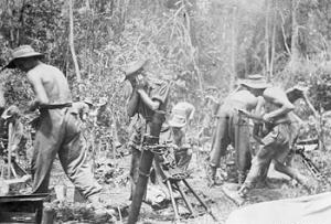 black and white photo\' of four men, three of them stripped to the waist, wearing baggy trousers and bush hats, setting up some sort of light artillery, with several other similar men and the beginnings of jungle in the background