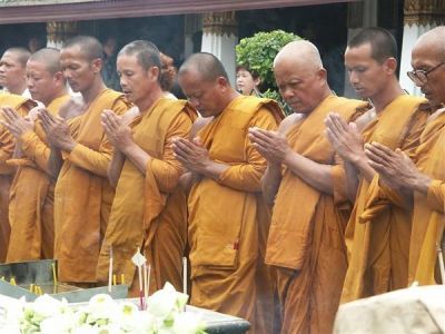 colour photo\' showing a row of eight men in yellow robes, with bare right shoulders and stoles draped over their left shoulders, standing with hands joined in prayer in front of a table bearing candles and possibly white flowers